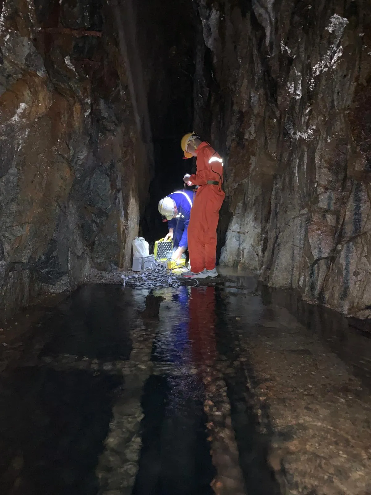 Two people in helmets preparing research equipment beside water inside a rocky mine passage.